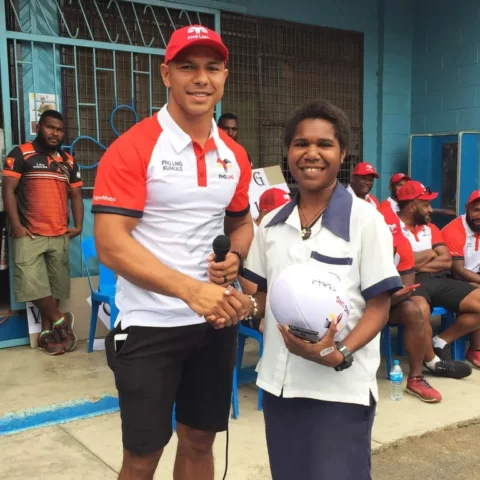 David Mead smiling shaking hand of student while doing community work with PNG Kumuls
